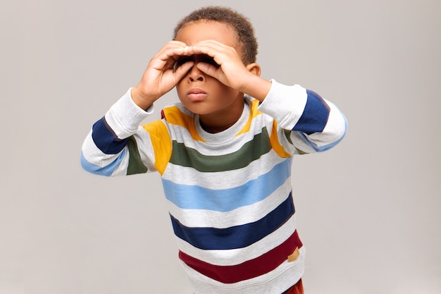 Geïsoleerde shot van nieuwsgierige afro-amerikaanse jongen die gebaar met beide handen in de buurt van de ogen maakt en door gaten kijkt alsof hij een verrekijker gebruikt, iets op afstand zoeken. jeugd en leuk concept
