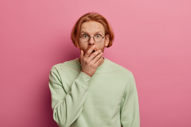 Geïsoleerde shot van verbijsterde bebaarde roodharige hipster man bedekt geopende mond, kan niet geloven in schokkend nieuws, ziet iets ongelooflijks, gekleed in casual trui, poseert tegen roze muur