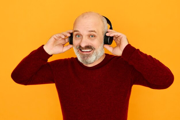 Geïsoleerde shot van vrolijke knappe blanke senior man met kaal hoofd en grijze baard glimlachend overnemen van moderne draadloze verbinding met elektronische gadget via bluetooth.