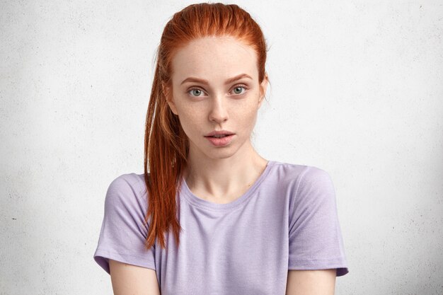 Geïsoleerde studio shot van mooie jonge vrouw heeft sproeten huid, rood haar, draagt casual paars t-shirt, kijkt serieus naar de camera, luistert aandachtig naar de gesprekspartner