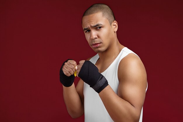 Geïsoleerde studio shot van woedende jonge Afrio Amerikaanse mannelijke vechter die wit mouwloos sportshirt en zwarte boksbandages draagt, camera kijkt met een felle blik tijdens het trainen in de sportschool