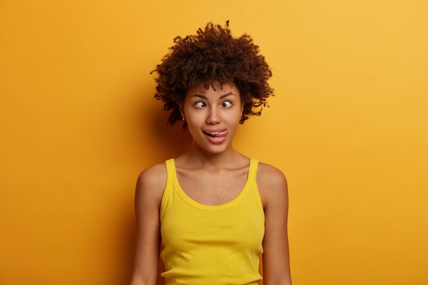 Gratis foto gekke afro-amerikaanse vrouw dwaas rond, trekt een grappig gezicht, kruist de ogen en steekt tong uit, toont grimas, voelt zich speels en gek, draagt een casual vest, staat tegen felgele muur.