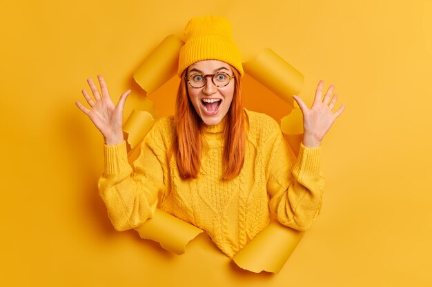 Gekke emotionele roodharige tienermeisje houdt mond wijd open steekt handpalmen op reageert op geweldig nieuws of grote uitverkoop draagt gele trui en hoed.