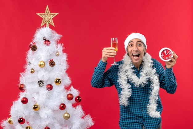 Gekke grappige jonge man met kerstman hoed en het verhogen van een glas wijn en klok in de buurt van kerstboom op rood