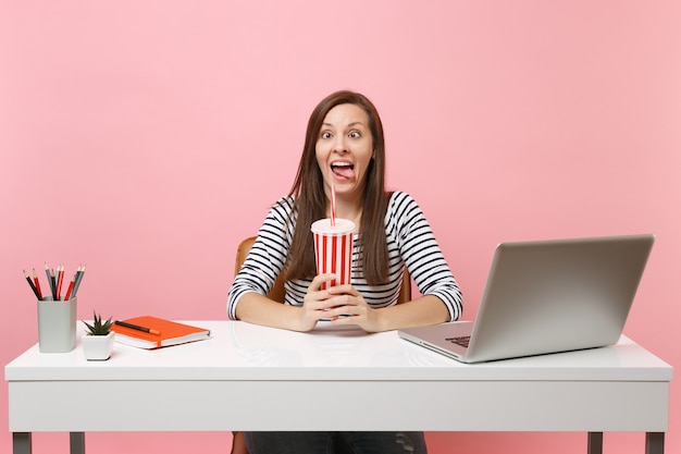 Gekke grappige vrouw die gezichten maakt met tongenknijpende ogen die een plastic beker met cola-frisdrank vasthouden aan een wit bureau met pc-laptop