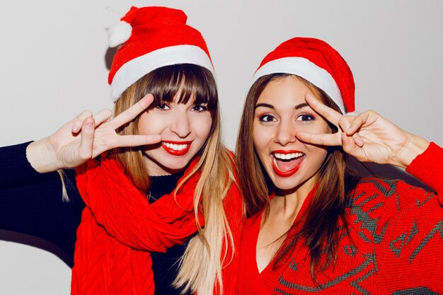 Gekke sfeer voor een nieuwjaarsfeest. Twee dronken lachende vrouwen met plezier en poseren in schattige maskeradehoeden. Rode trui en sjaal. Tekenen tonen. Witte tanden, lichte make-up.