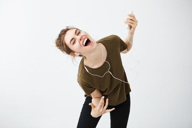 Gekke vrolijke jonge vrouw die zingende het luisteren muziek in hoofdtelefoons over witte achtergrond dansen.