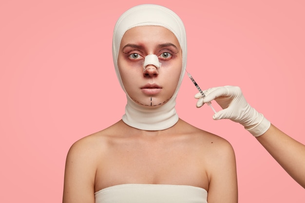 Gekneusde jonge vrouw in verband krijgt injectie in gezichtszone, vult gezicht met collageen, onderging ooglidcorrectie, neuscorrectie en kinreductie