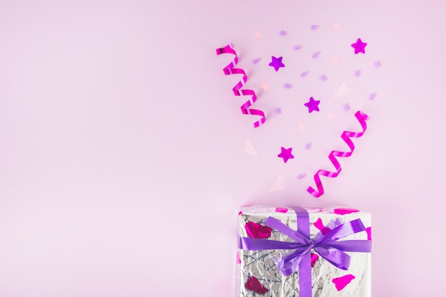 Gekruld slingers, stervorm en confetti over de zilveren geschenkdoos tegen roze achtergrond