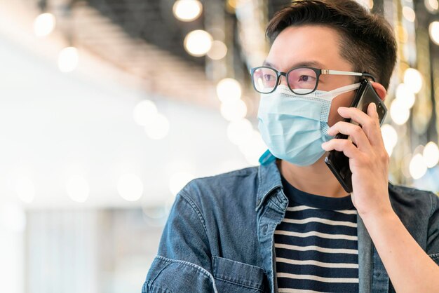 Geluk aziatische volwassen volwassen man die een beschermend gezichtsmasker draagt, gebruik smartphone-apparaatcommunicatie met de achtergrond van wazig bokeh-licht in supermarkt-winkelcentrum