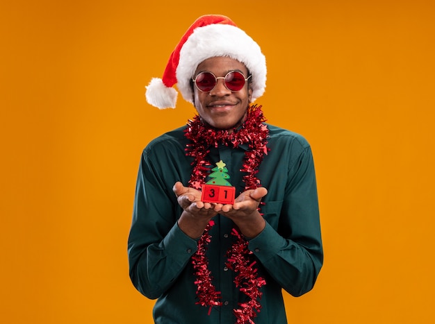 Gelukkig Afro-Amerikaanse man in KERSTMUTS met garland dragen van een zonnebril speelgoed kubussen met Nieuwjaar datum kijken camera glimlachend vrolijk permanent over oranje achtergrond