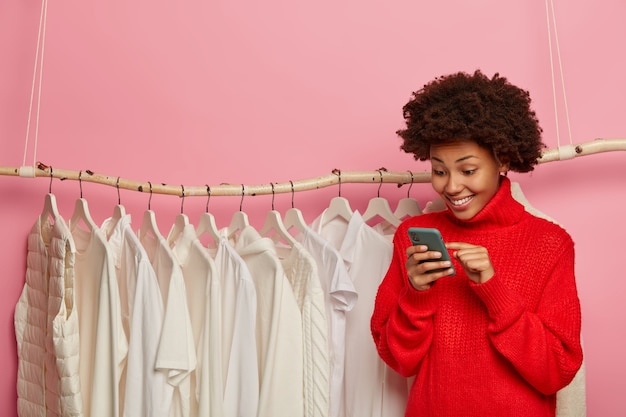 Gelukkig afro-amerikaanse manager van online winkel, raadpleegt klanten, typeert commentaar, verkoopt nieuwe collectie in witte kleur, draagt gebreide rode trui, staat in de buurt van rek, geïsoleerd over roze muur.