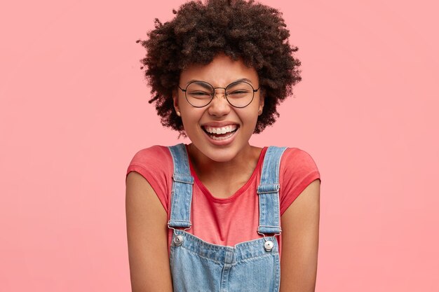 Gelukkig Afro-Amerikaanse vrouw giechelt en sluit de ogen, haalt haar schouders op, gekleed in een casual t-shirt en denim tuinbroek, poseert tegen roze muur. Mensen, positiviteit, emoties concept.