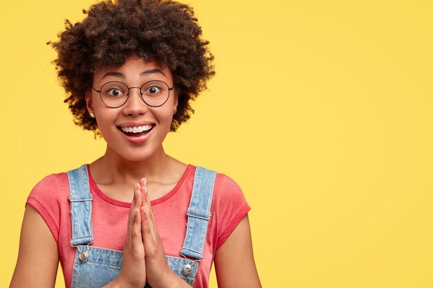 Gratis foto gelukkig afro-amerikaanse vrouw met brede glimlach, houdt de handpalmen tegen elkaar gedrukt, gelooft in wonder, gekleed in roze t-shirt en overall, poseert over gele muur met vrije ruimte aan de rechterkant