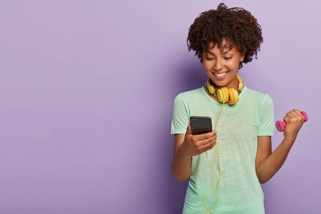 Gelukkig atleet vrouwtje heeft afro kapsel, treinen met halter, luistert naar muziek via koptelefoon, kijkt naar telefoon, gekleed in casual t-shirt