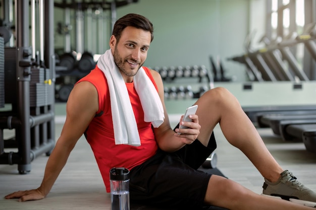 Gelukkig atletische man met behulp van mobiele telefoon terwijl u ontspant na sporttraining in een sportschool