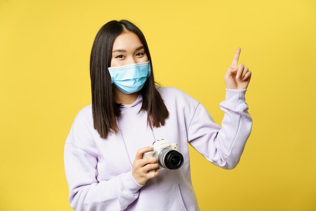 Gelukkig Aziatisch meisje met gezichtsmasker, foto's maken, vinger wijzen naar kopieerruimte, camera vasthouden, over gele achtergrond staan.