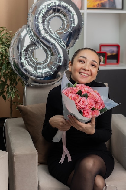 Gelukkig Aziatische jonge vrouw in zwarte jurk zittend op een stoel met een boeket bloemen glimlachend vrolijk in lichte woonkamer internationale Vrouwendag vieren