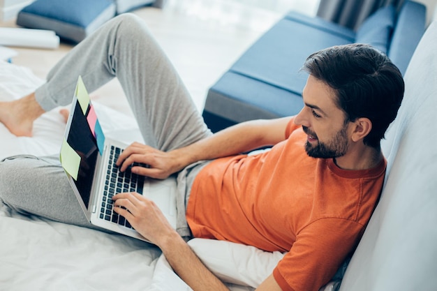 Gelukkig bebaarde jonge man zittend op een comfortabel bed en typen tijdens het gebruik van een laptop
