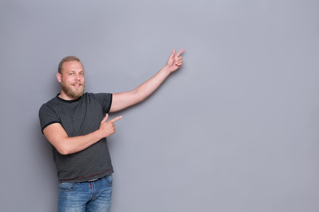 Gelukkig bebaarde man ergens wijzen. Knappe volwassen man in zwart T-shirt glimlachend voor de camera op grijs.