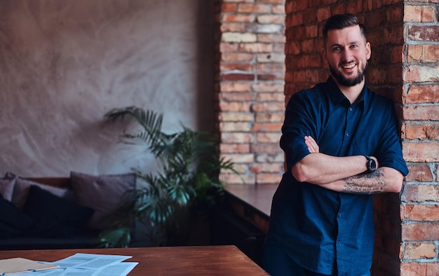 Gratis foto gelukkig bebaarde man permanent met gekruiste armen in de buurt van desktop in kantoor met loft interieur.