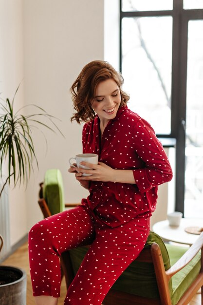 Gelukkig blanke vrouw in pyjama genieten van ochtend. Binnen schot van innemende krullende vrouw met een kopje koffie.