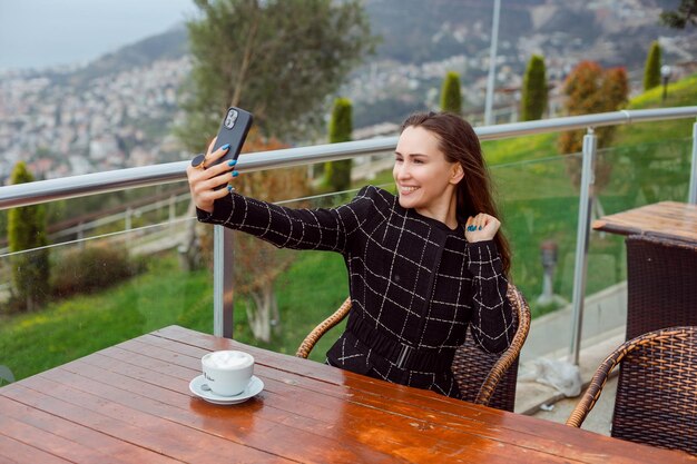 Gelukkig bloggermeisje neemt selfie met haar smartphone door hand op te steken door op de achtergrond van de stad te zitten