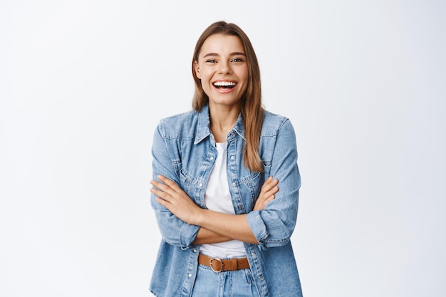 Gelukkig blond meisje in vrijetijdskleding, lachend om een grappige grap, er zorgeloos uitzien en plezier hebben, staande met gekruiste armen op de borst, wit