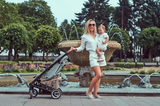 Gratis foto gelukkig blonde vrouw gekleed in een witte blouse, korte broek en zonnebril houdt haar dochtertje op handen, poseren in de buurt van een fontein in het park.