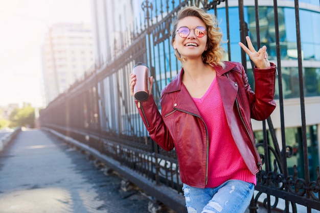 Gelukkig blonde vrouw poseren op moderne straten, koffie drinken of cappuccino. Stijlvolle herfstoutfit, leren jas en gebreide trui. Roze zonnebril.