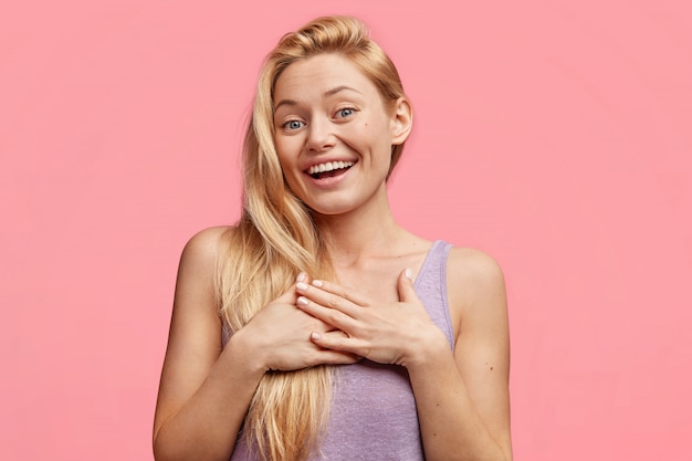 Gelukkig blonde vrouw wordt aangeraakt en blij om gefeliciteerd te ontvangen van vrienden, houdt de handen op de borst, lacht aangenaam, nonchalant gekleed