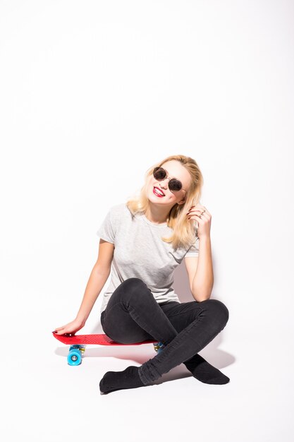 Gelukkig Blondie met gekruiste benen zit op rode skateboard voor witte muur