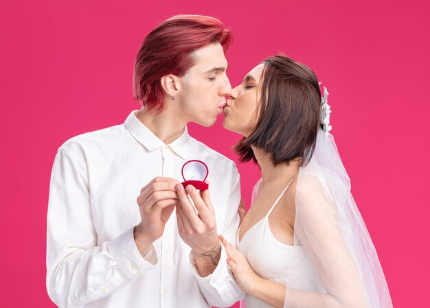 Gelukkig bruidspaar van een man die een huwelijksaanzoek doet met een trouwring in een geschenkdoos, gelukkig verliefd samen zoenen over roze muur