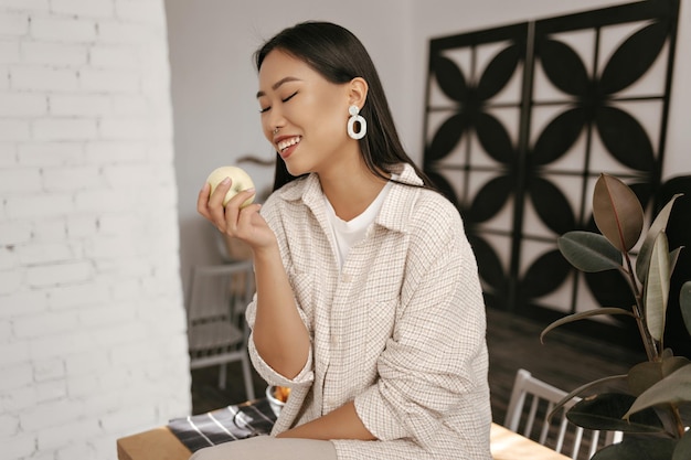 Gelukkig brunette aziatische dame in beige vest en broek glimlacht oprecht en kijkt naar verse appel cool gebruinde vrouw zit op houten tafel op keuken en houdt fruit