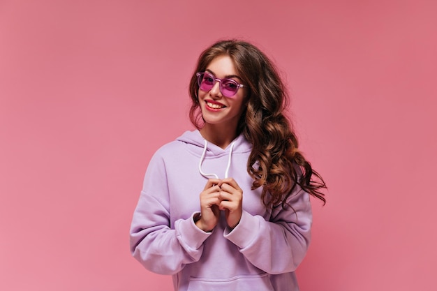 Gelukkig brunette krullend vrouw in paarse oversized hoodie kijkt naar de camera Meisje in heldere zonnebril glimlacht op geïsoleerde roze achtergrond