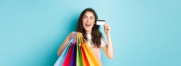 Gelukkig brunette meisje shopaholic die haar plastic creditcard en boodschappentassen toont, kijkt dromerig naar het logo of de banner van de winkel die over een blauwe achtergrond staat