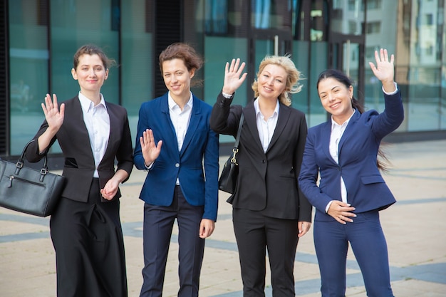 Gelukkig business damesteam hallo zwaaien, samen in de buurt van kantoorgebouw, camera kijken en glimlachen. Medium shot, vooraanzicht. Zakenvrouwen groepsportret concept