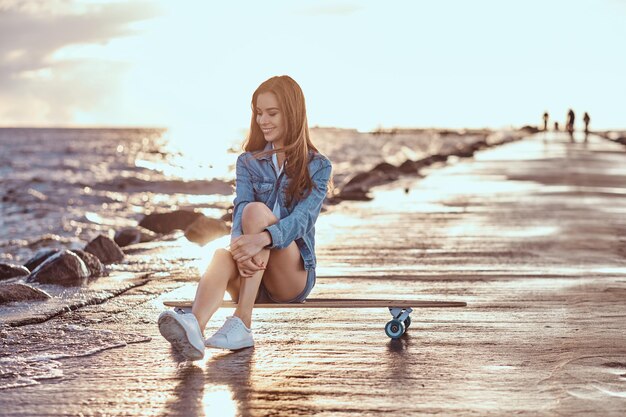Gelukkig charmant meisje gekleed in korte broek en een t-shirt zit op een skateboard op het strand tijdens heldere zonsondergang.