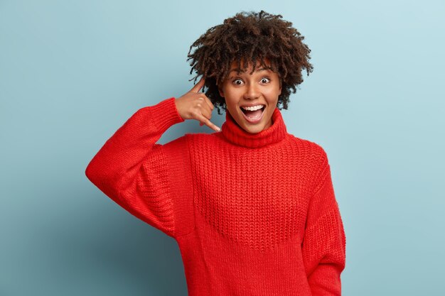 Gelukkig dolgelukkig Afro-Amerikaanse vrouw toont bel me gebaar, vraagt contact houden, draagt warme rode trui, in hoge geest, geïsoleerd over blauwe muur. Mensen en lichaamstaal concept.