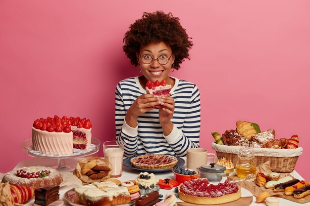 Gelukkig donkere vrouw eet smakelijke aardbeientaart, draagt gestreepte trui, poseert aan tafel overladen met desserts, krijgt veel plezier, poseert over roze muur