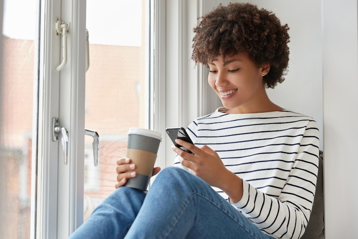 Gratis Foto gelukkig donkere vrouw met tevreden uitdrukking, leest online boek via mobiele telefoon, controleert e-mail in sociale netwerken