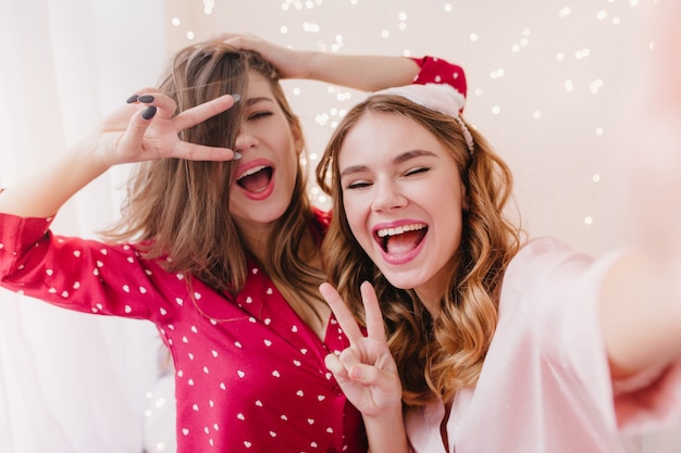 Gelukkig donkerharige meisje gek rond tijdens ochtendfotoshoot. Charmante vrouw lachen in roze pyjama selfie met vriend maken.