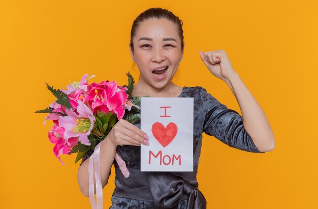 Gelukkig en opgewonden Aziatische vrouw met wenskaart en boeket bloemen vieren internationale Vrouwendag balde vuist staande over oranje muur