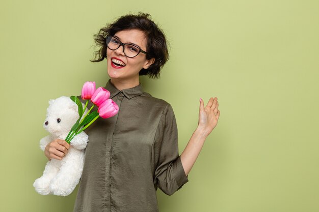 Gelukkig en positieve vrouw met kort haar met een boeket tulpen en teddybeer camera kijken glimlachend vrolijk zwaaien met hand vieren internationale vrouwendag 8 maart