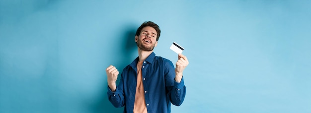 Gelukkig en tevreden jonge man dansen met plastic creditcard vuist pomp en glimlach staande op blauw