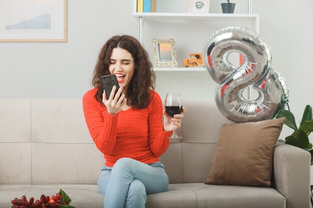 Gelukkig en tevreden jonge vrouw in casual kleding glimlachend vrolijk zittend op een bank met glas wijn praten op mobiele telefoon in lichte woonkamer vieren internationale vrouwendag 8 maart