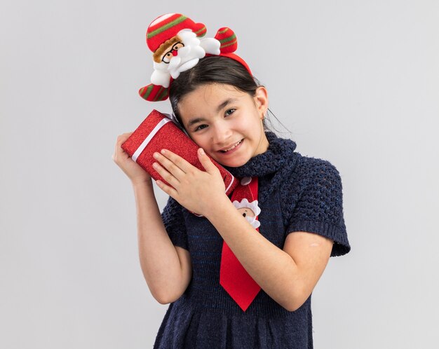 Gelukkig en tevreden meisje in gebreide jurk die rode stropdas met grappige kerstrand op hoofd bedrijf kerstcadeau op zoek glimlachen