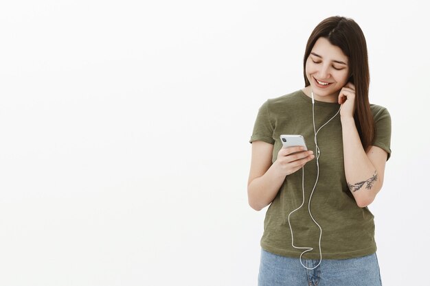 Gelukkig en tevreden vrolijk jong meisje oortelefoon in oor zetten als kantelend hoofd en neerkijken op smartphonescherm met tedere glimlach willen luisteren nieuw geweldig liedje, nummer kiezen in app