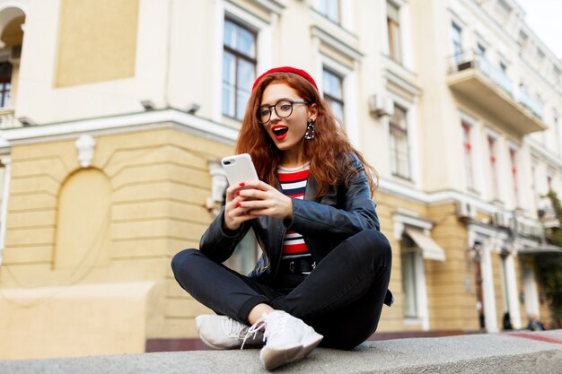 Gelukkig fantastische gember vrouw in stijlvolle rode baret in de straat met behulp van haar smartphone