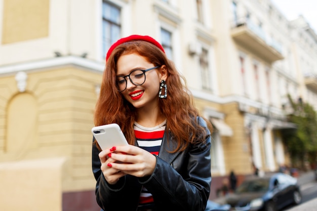 Gelukkig fantastische gember vrouw in stijlvolle rode baret in de straat met smartphone